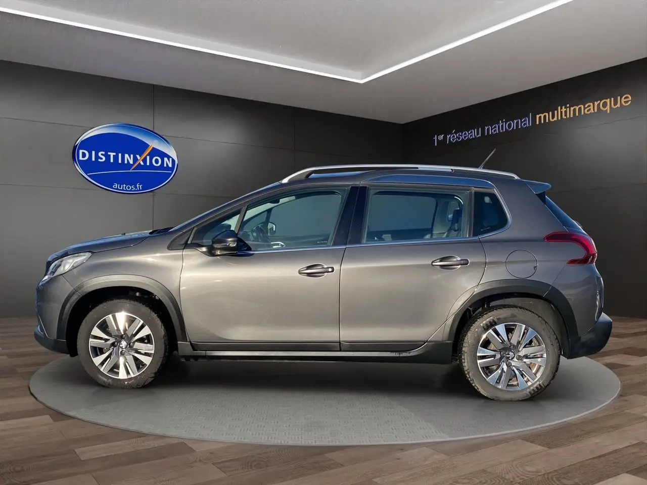 Vue de profil côté gauche d'un Peugeot 2008 gris platinium avec jantes alliage et barres de toit en intérieur showroom.