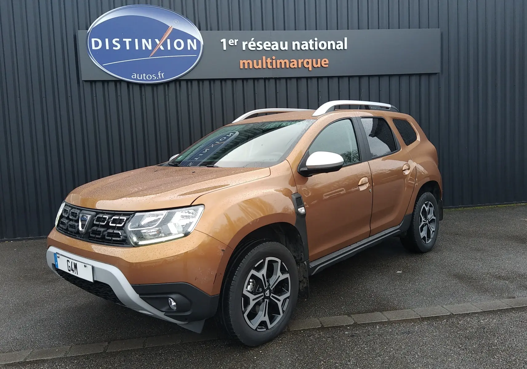 Dacia Duster orange Atacama en 3/4 avant droit, avec jantes alliage et barres de toit blanches visibles.