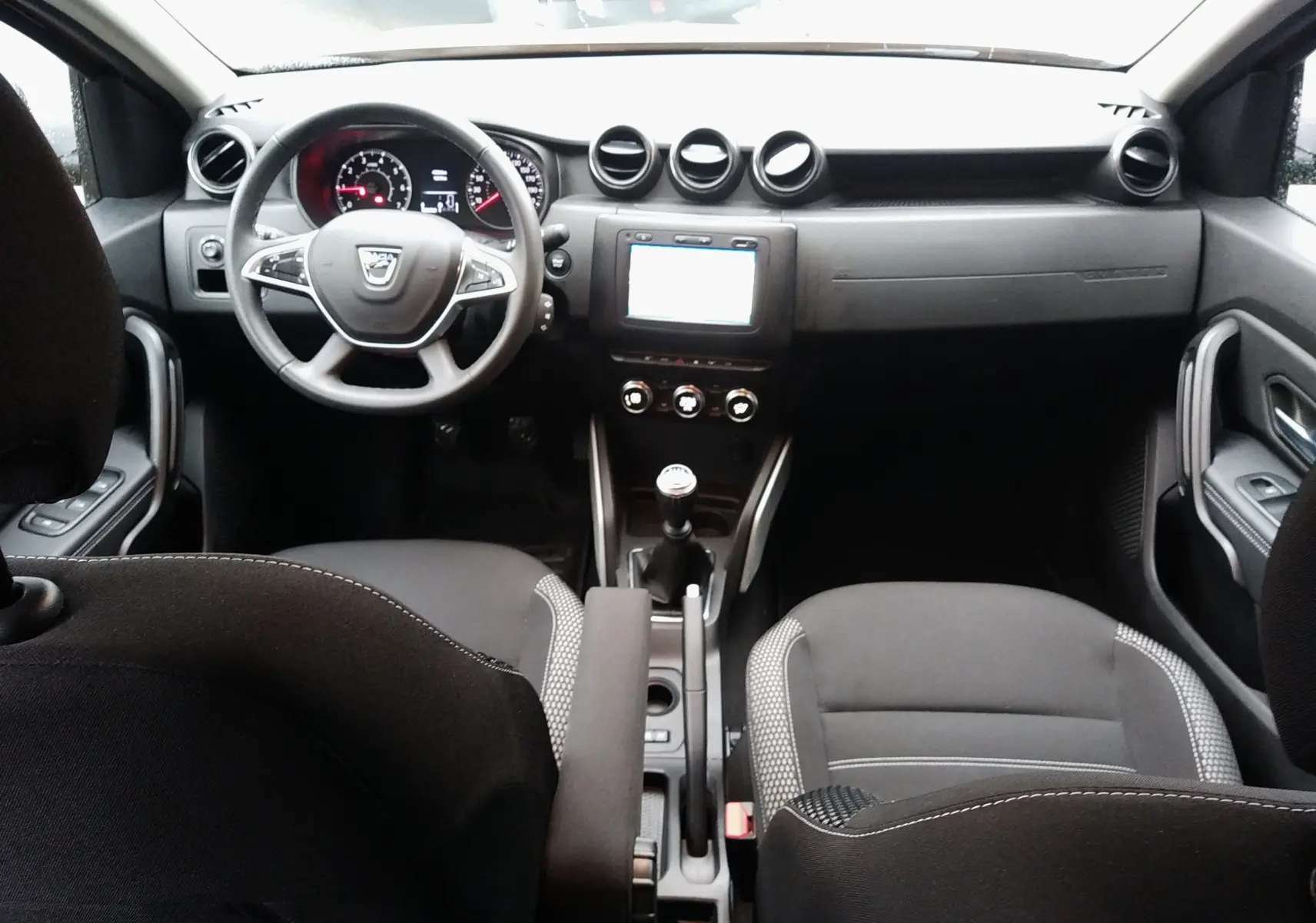 Vue intérieure avant du Dacia Duster 2021, tableau de bord et volant noirs avec console centrale et levier de vitesses manuelle.