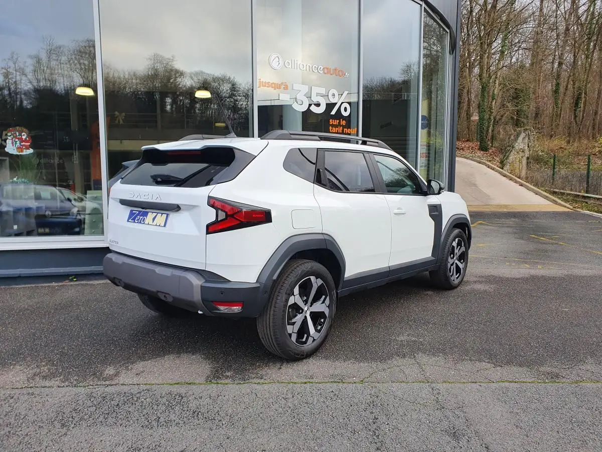 Vue 3/4 arrière droite du Dacia Duster blanc glacier 2025 avec jantes alliage 18” et barres de toit longitudinales.