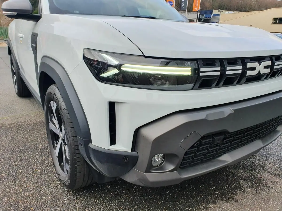 Vue 3/4 avant gauche du Dacia Duster blanc glacier 2025 avec calandre noire et phares LED allumés.