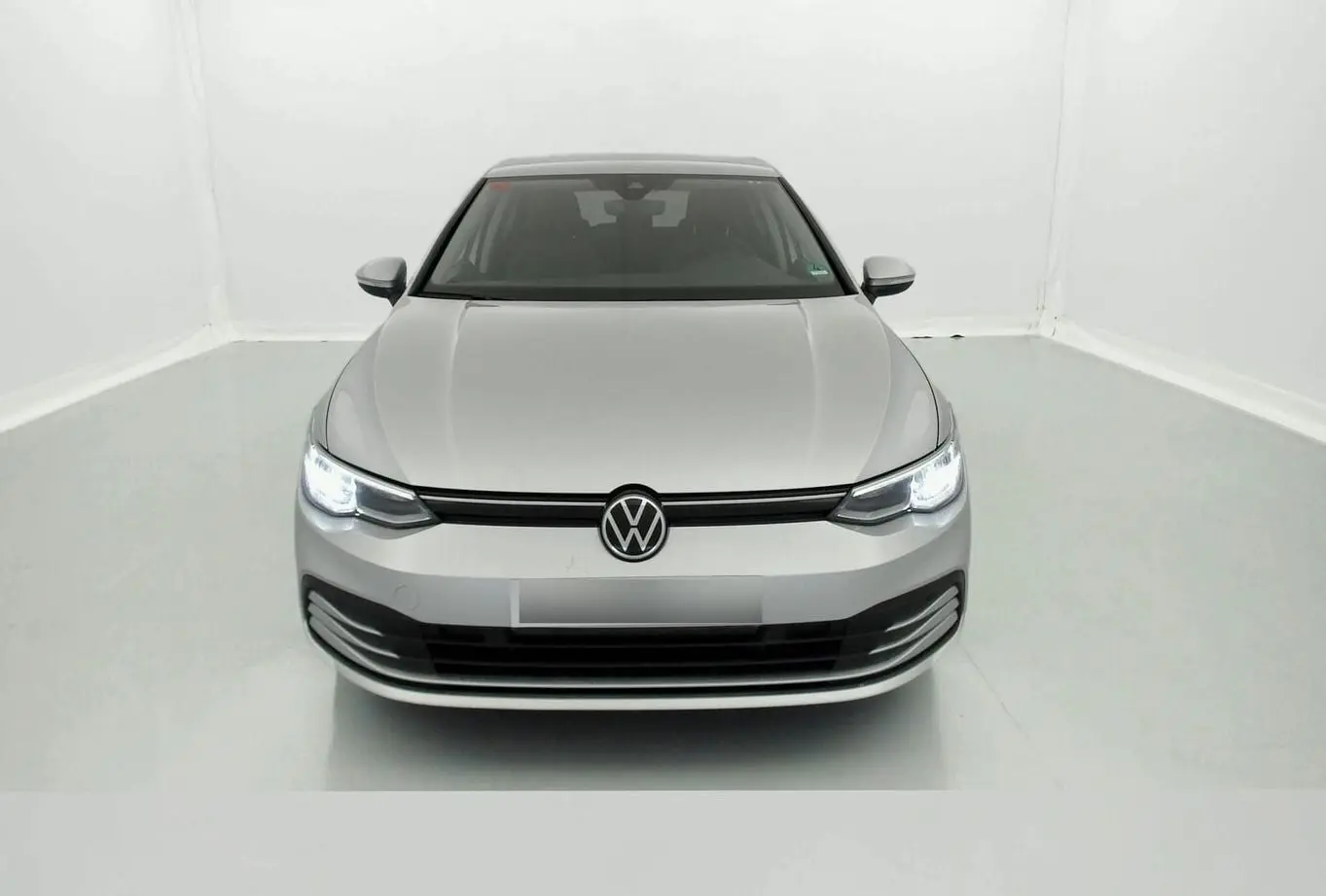 Vue frontale d'une Volkswagen Golf VIII gris reflex avec phares allumés dans un studio blanc.