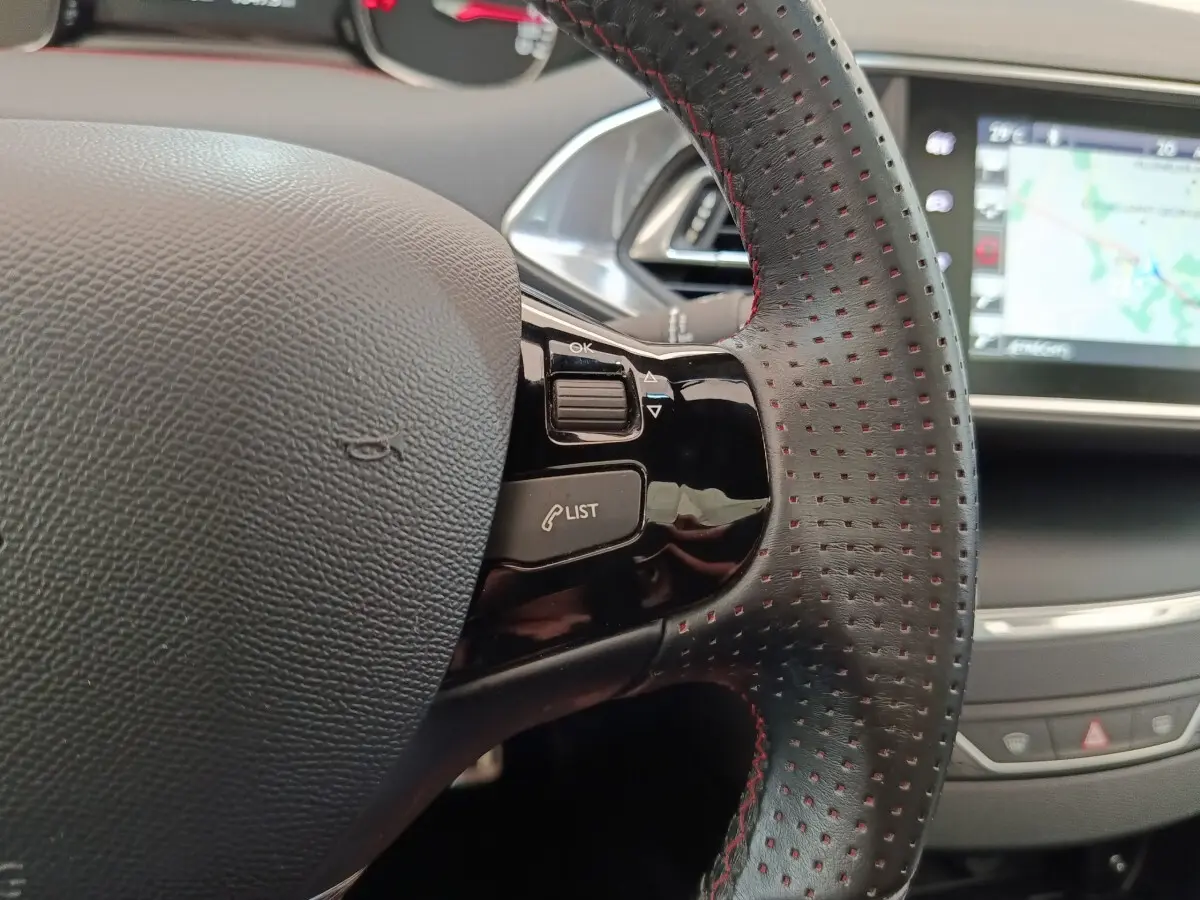 Gros plan sur le volant cuir noir perforé avec surpiqûres rouges de la Peugeot 308, écran tactile de navigation en arrière-plan.
