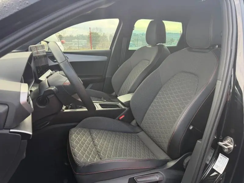 Vue intérieure côté conducteur de la SEAT LEON ST 1.5 eTSI Hybrid 2025, sièges tissu noir avec surpiqûres rouges.