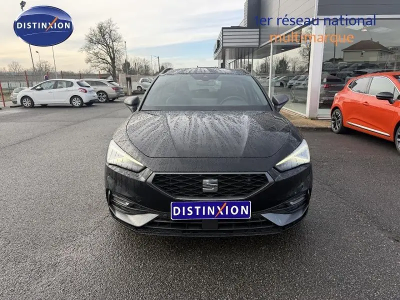 Vue frontale d'une SEAT LEON ST 1.5 eTSI Hybrid noire métallisée, avec calandre hexagonale et phares allumés sous la pluie.