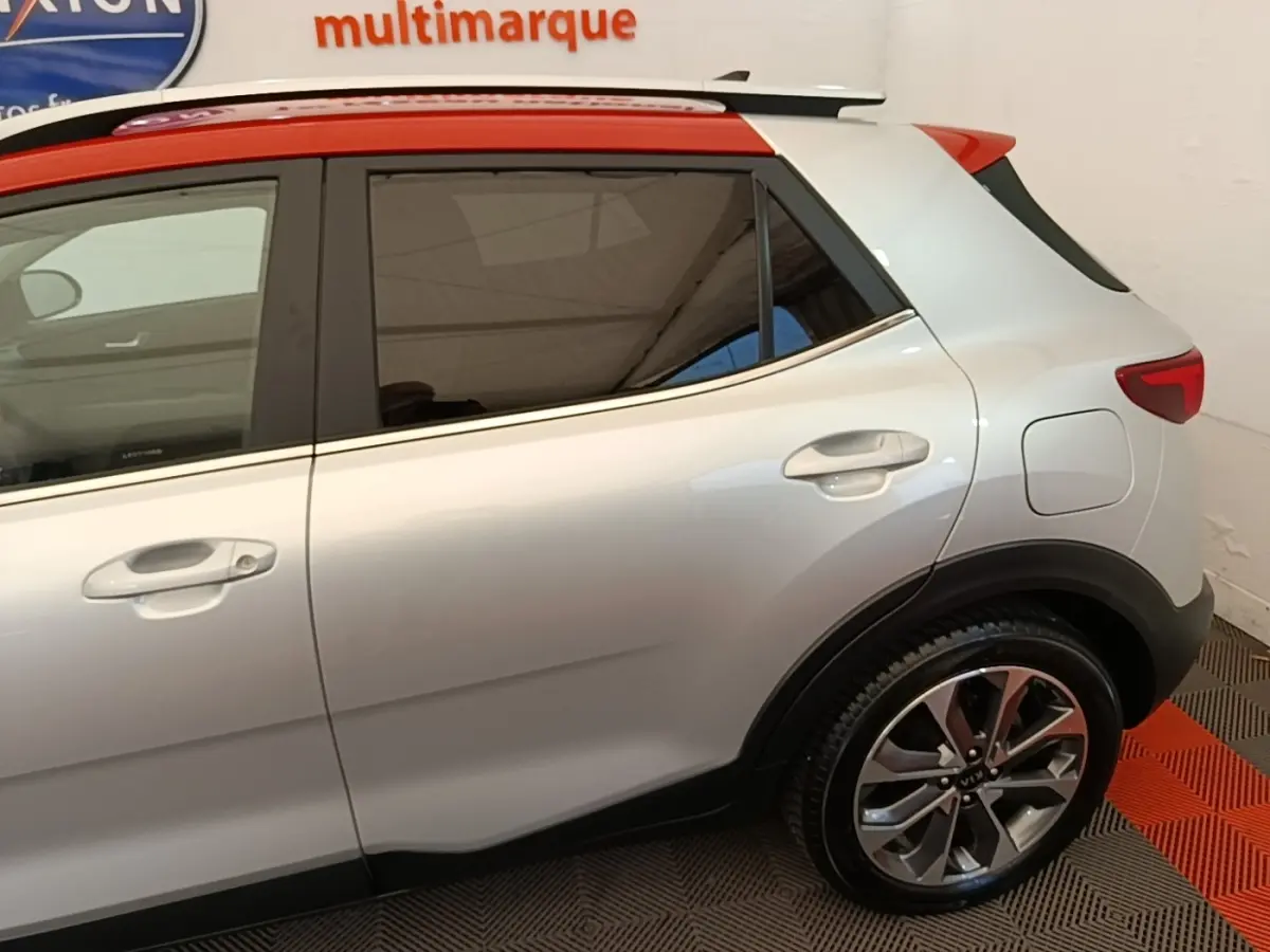 Vue latérale droite d'un KIA Stonic gris clair avec toit rouge et jantes alliage, en intérieur showroom.