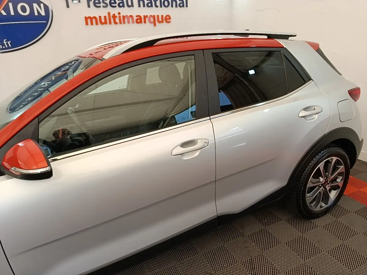 KIA Stonic gris clair vue de profil côté gauche, toit et rétroviseur orange, jantes alliage et rails de toit chromés.