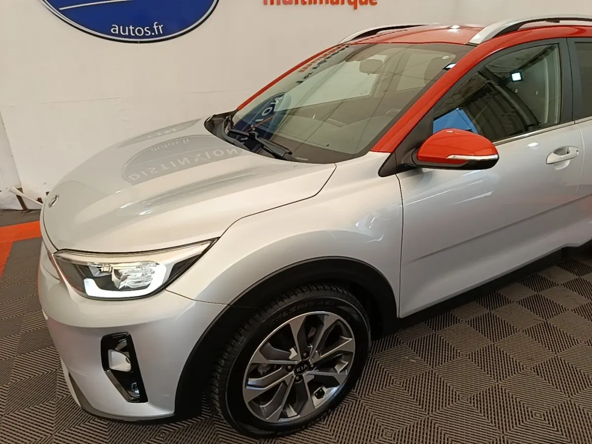 Vue 3/4 avant droit d’un KIA Stonic gris clair avec toit et rétroviseur orange vif en intérieur showroom.