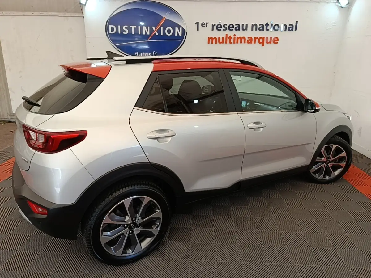 Vue 3/4 arrière droite d’un KIA Stonic gris clair avec toit rouge et jantes alliage, en intérieur showroom.