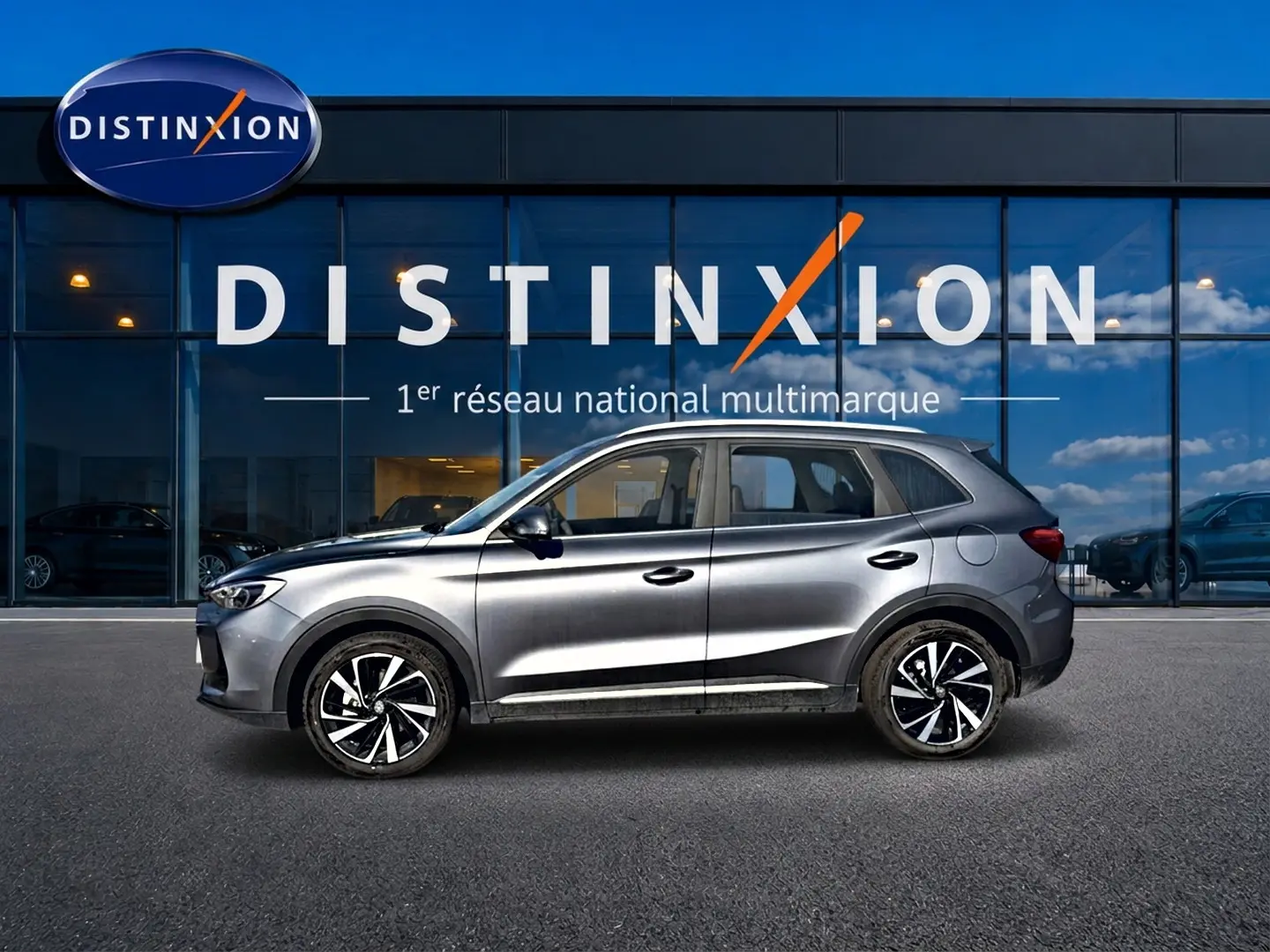 Vue latérale droite du MG Motor ZS gris 2026 avec jantes alliage noires et toit surélevé devant une vitrine Distinxion.
