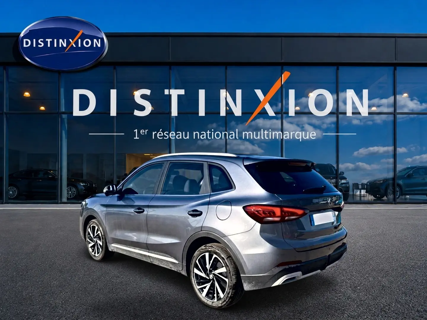 MG Motor ZS gris en 3/4 arrière droit, avec jantes alliage et feux arrière LED visibles devant un showroom Distinxion.