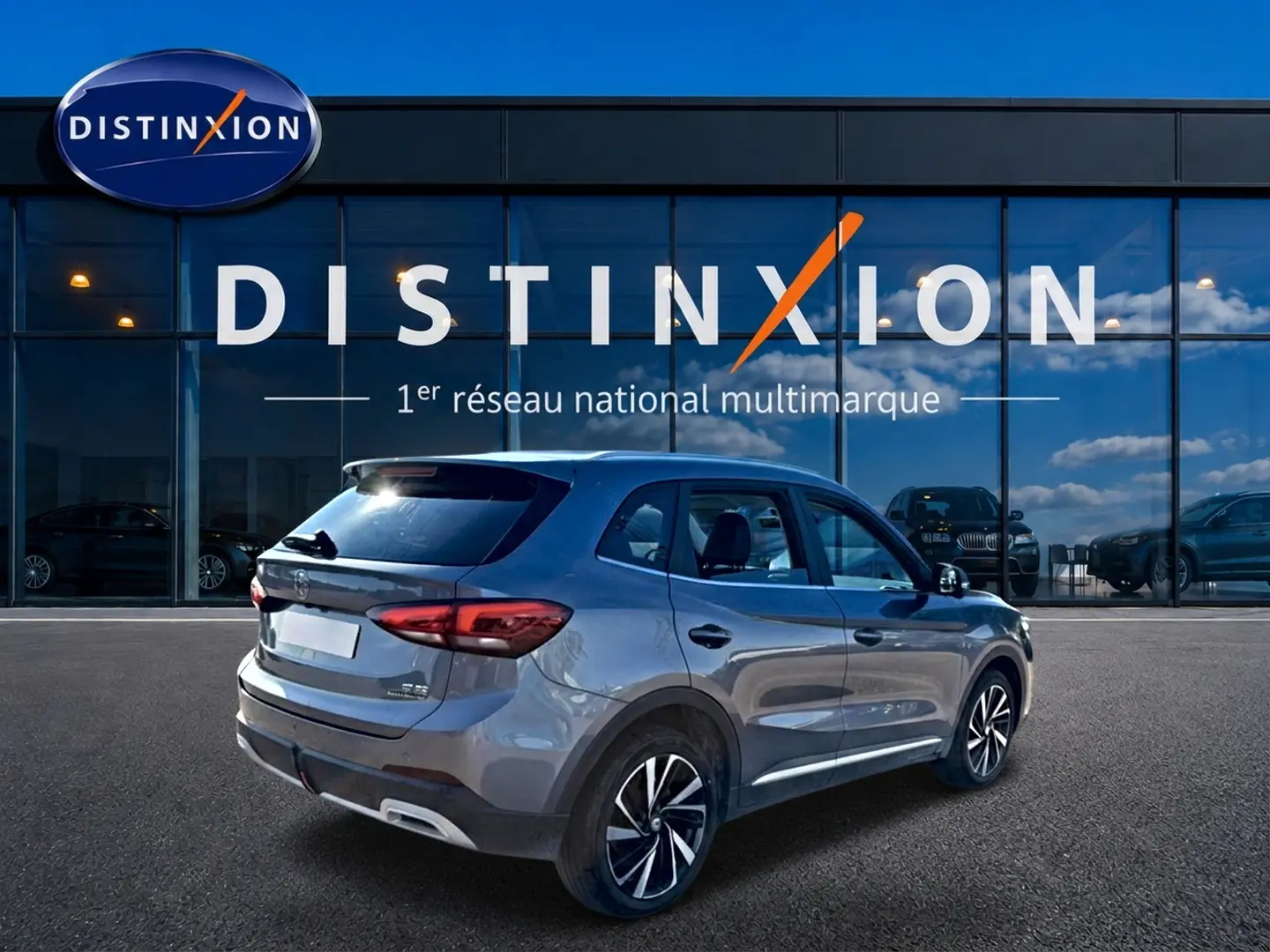 Vue 3/4 arrière droite d'un MG ZS gris 2026 avec jantes alliage noires et becquet arrière, devant un showroom Distinxion.