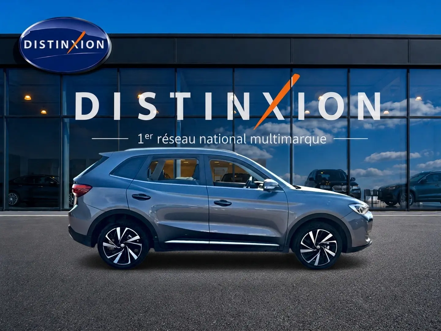 Vue de profil côté gauche du MG Motor ZS gris 1.5 hybrid 197 luxury devant une vitrine Distinxion.