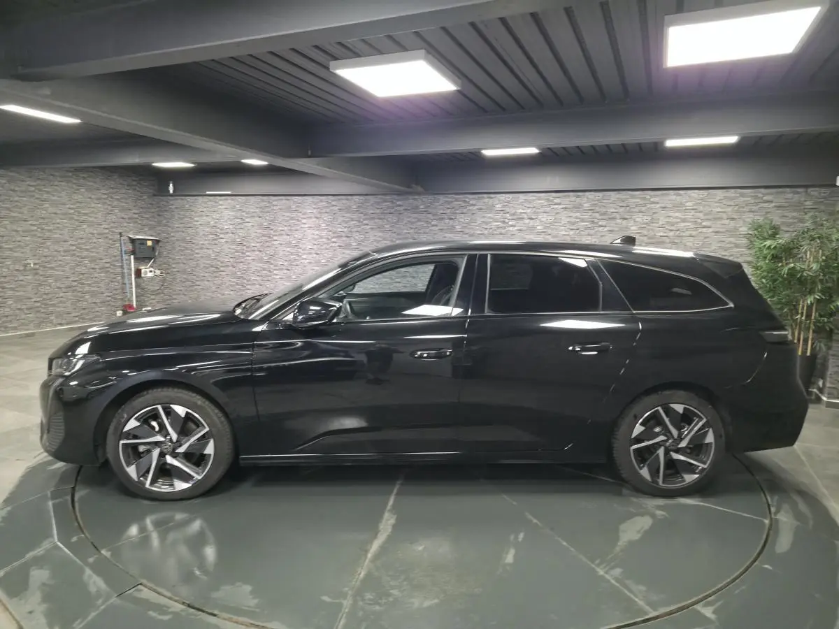 Peugeot 308 SW noire vue de profil côté gauche, jantes alliage 17'' et vitres surteintées en intérieur showroom.