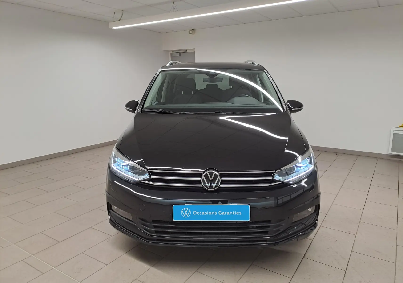 Vue avant d'un Volkswagen Touran 2025 noir intense nacré avec phares allumés dans un showroom.