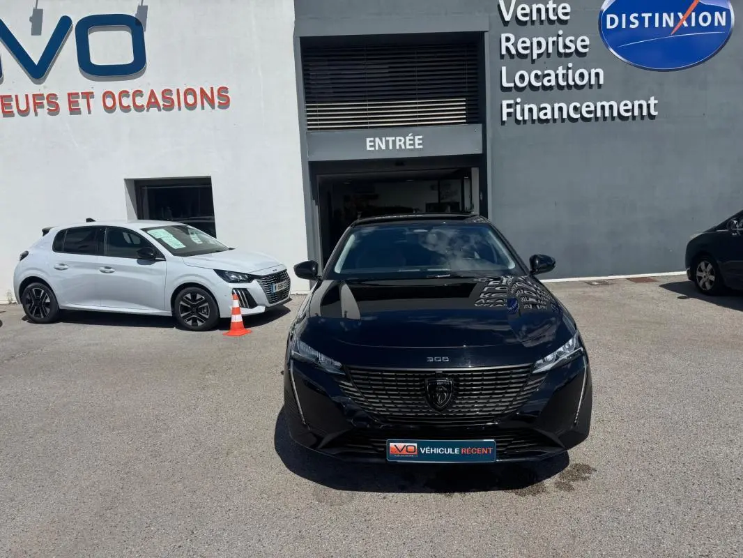 Vue de face d'une Peugeot 308 Hybrid 136 e-DCS6 Allure noire, stationnée devant un garage avec enseigne visible.
