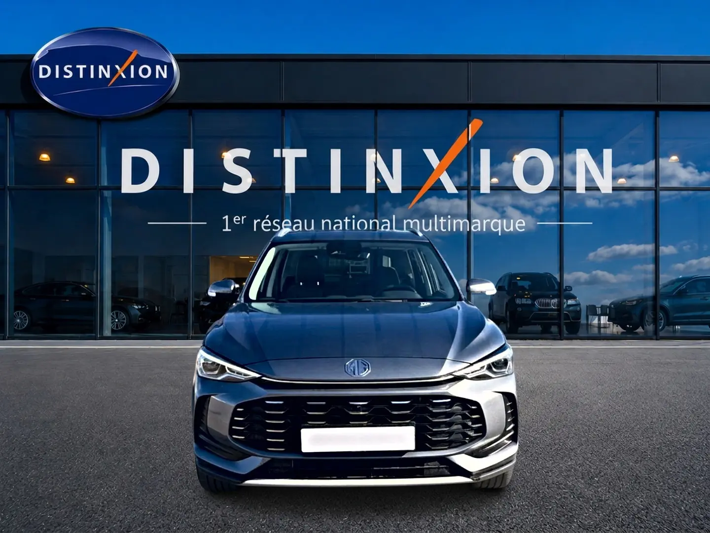 Vue frontale d'un MG MOTOR ZS 1.5 hybrid gris 2026 avec calandre noire et phares LED devant un showroom Distinxion.