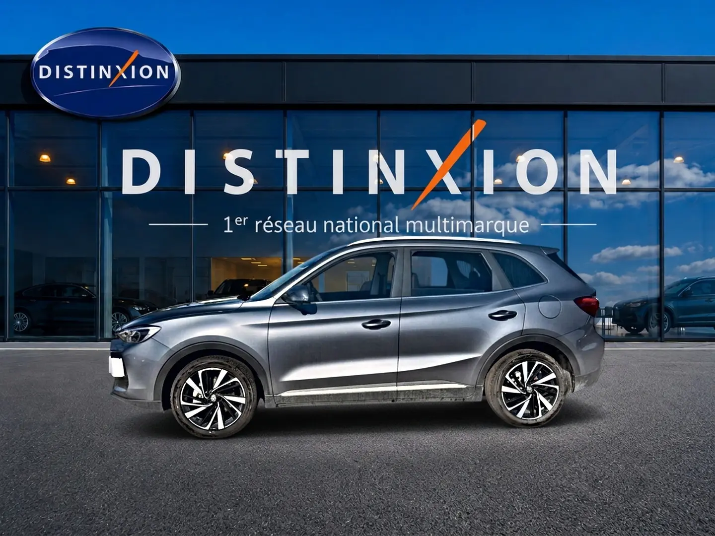 Vue de profil côté gauche d’un MG ZS gris 2026 hybride avec jantes alliage noires et blanches, devant une vitrine Distinxion.