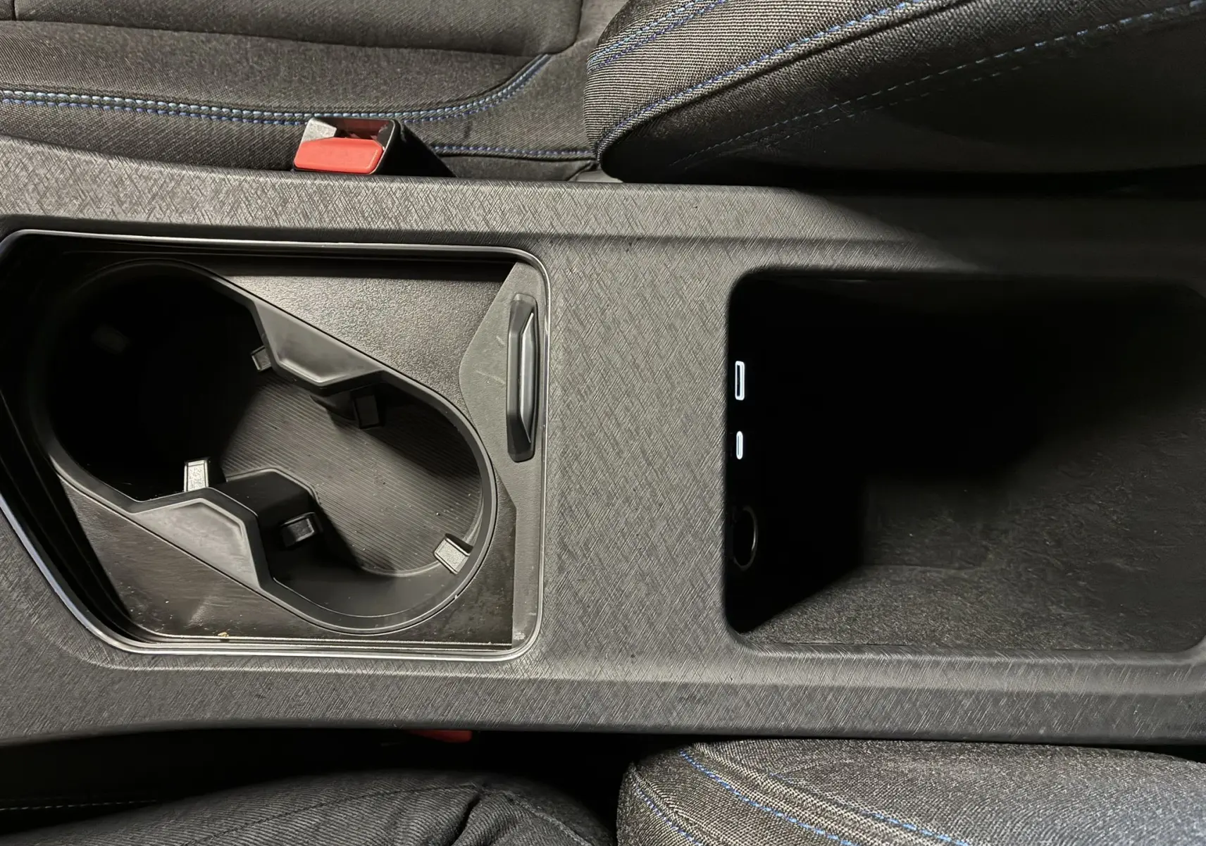 Gros plan sur la console centrale noire avec porte-gobelets et rangement, sièges tissu noir avec surpiqûres bleues.