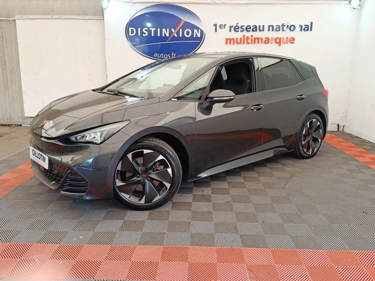 Vue 3/4 avant droite d'une CUPRA BORN gris foncé avec jantes noires et détails sportifs visibles en intérieur showroom.