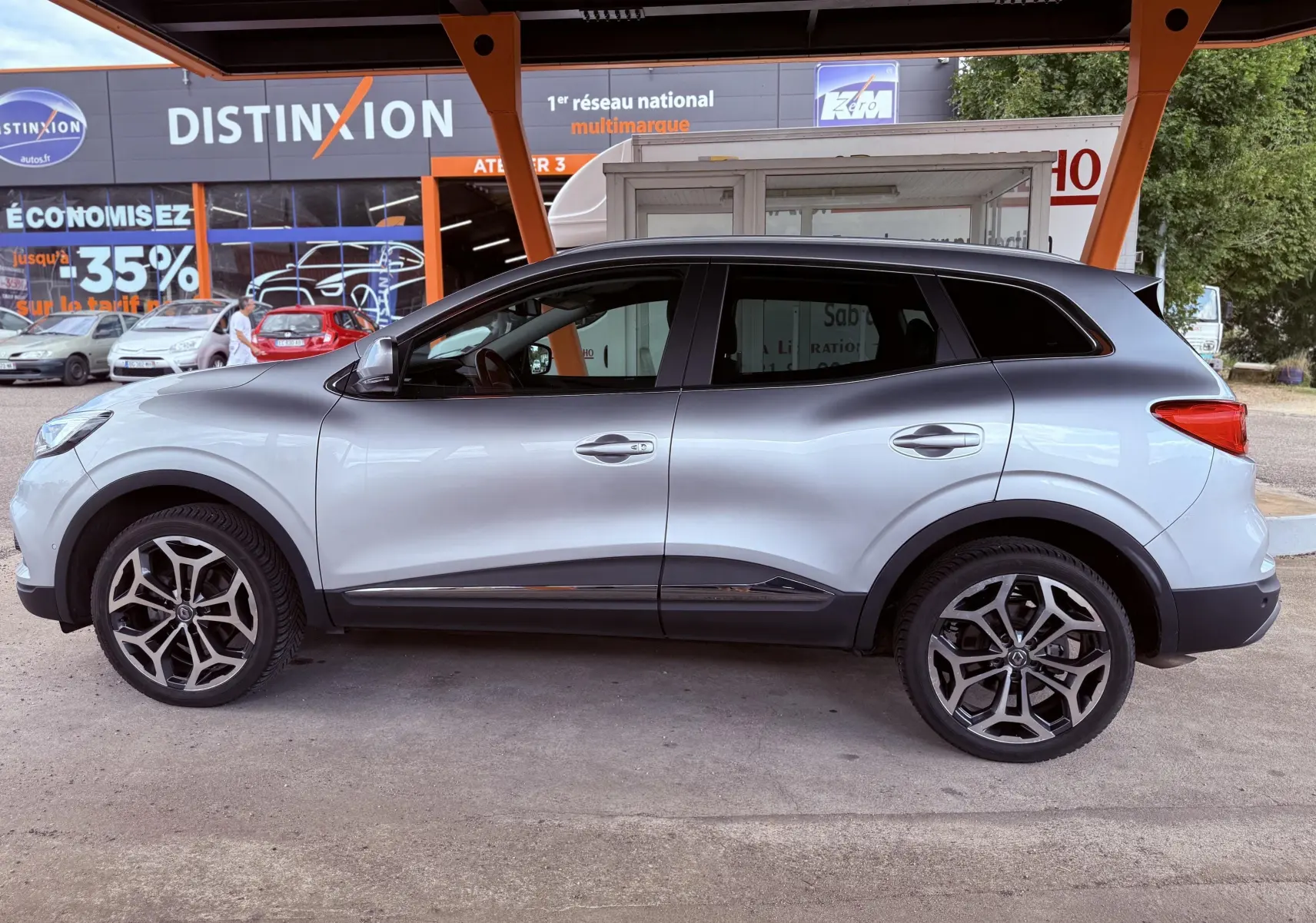 Profil côté gauche d'un Renault Kadjar gris 2020 avec jantes alliage et toit noir sous un abri commercial.