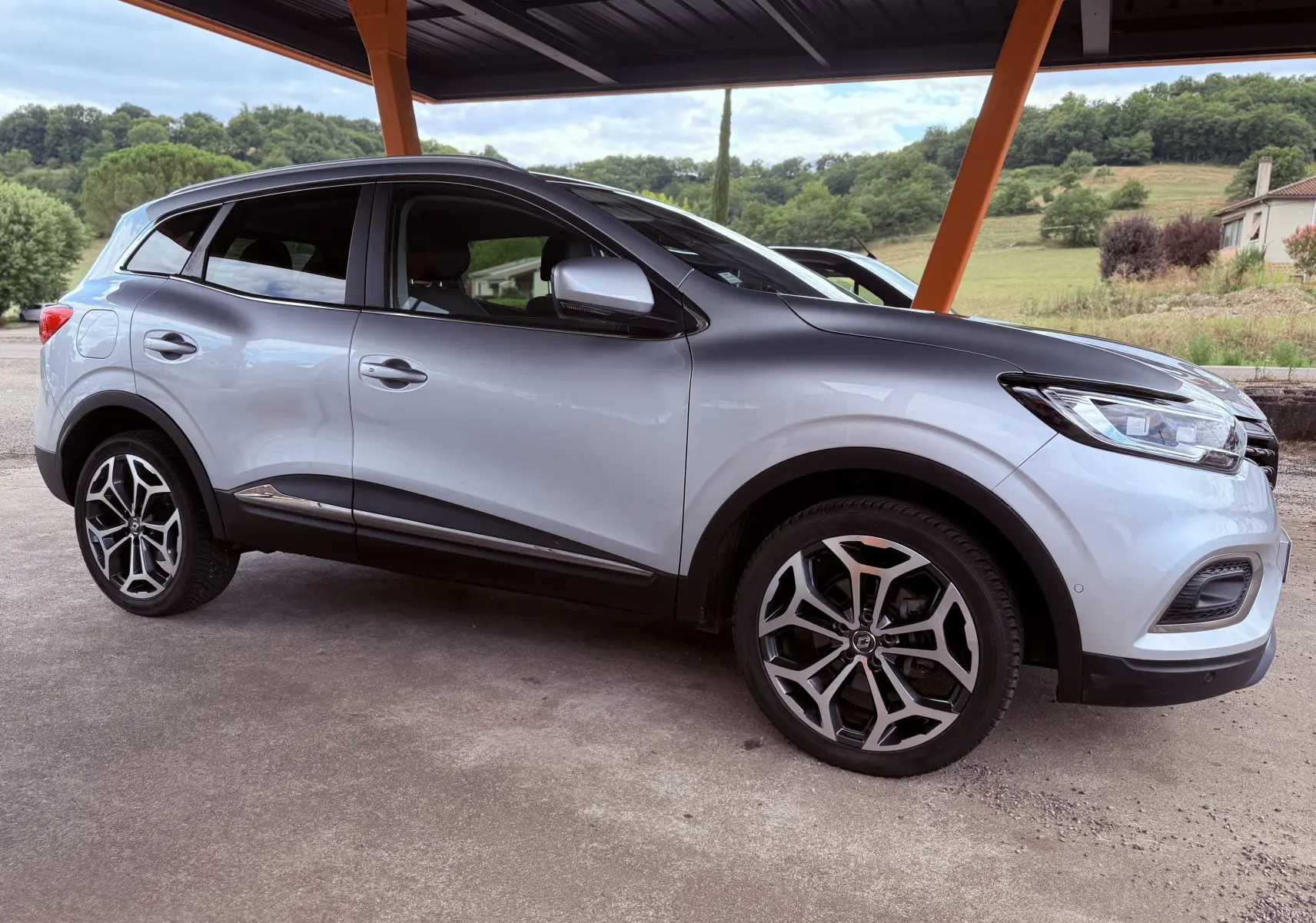 Renault Kadjar gris en vue de profil côté gauche, sous un abri avec paysage champêtre en arrière-plan.