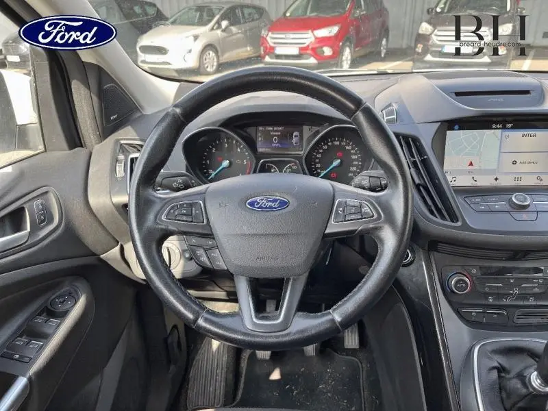 Vue intérieure centrée sur le volant cuir multifonction du Ford Kuga blanc glacier, avec tableau de bord et écran GPS visible.