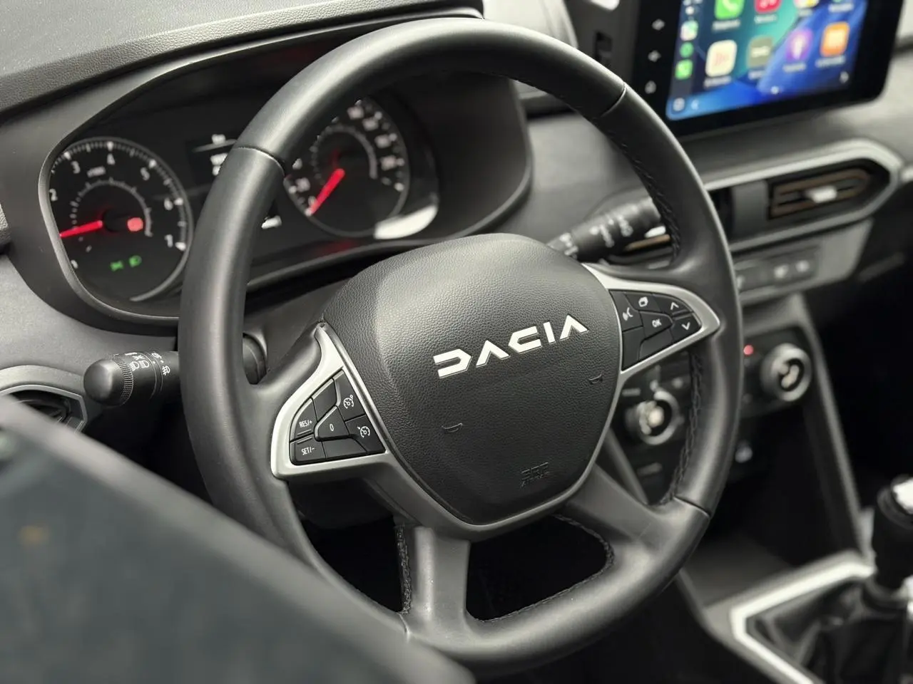 Vue rapprochée du volant noir Dacia avec commandes intégrées et écran tactile du tableau de bord en arrière-plan.