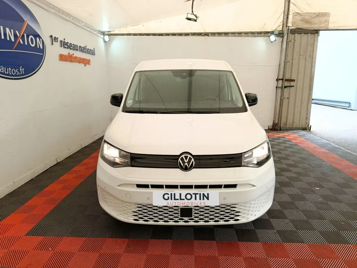 Vue avant d'un Volkswagen Caddy blanc 2022 avec calandre noire et logo VW centré, en intérieur sur sol damier rouge et noir.