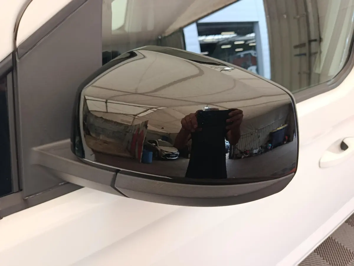 Rétroviseur extérieur droit noir brillant sur Volkswagen Caddy blanc, vue rapprochée en intérieur d'atelier.