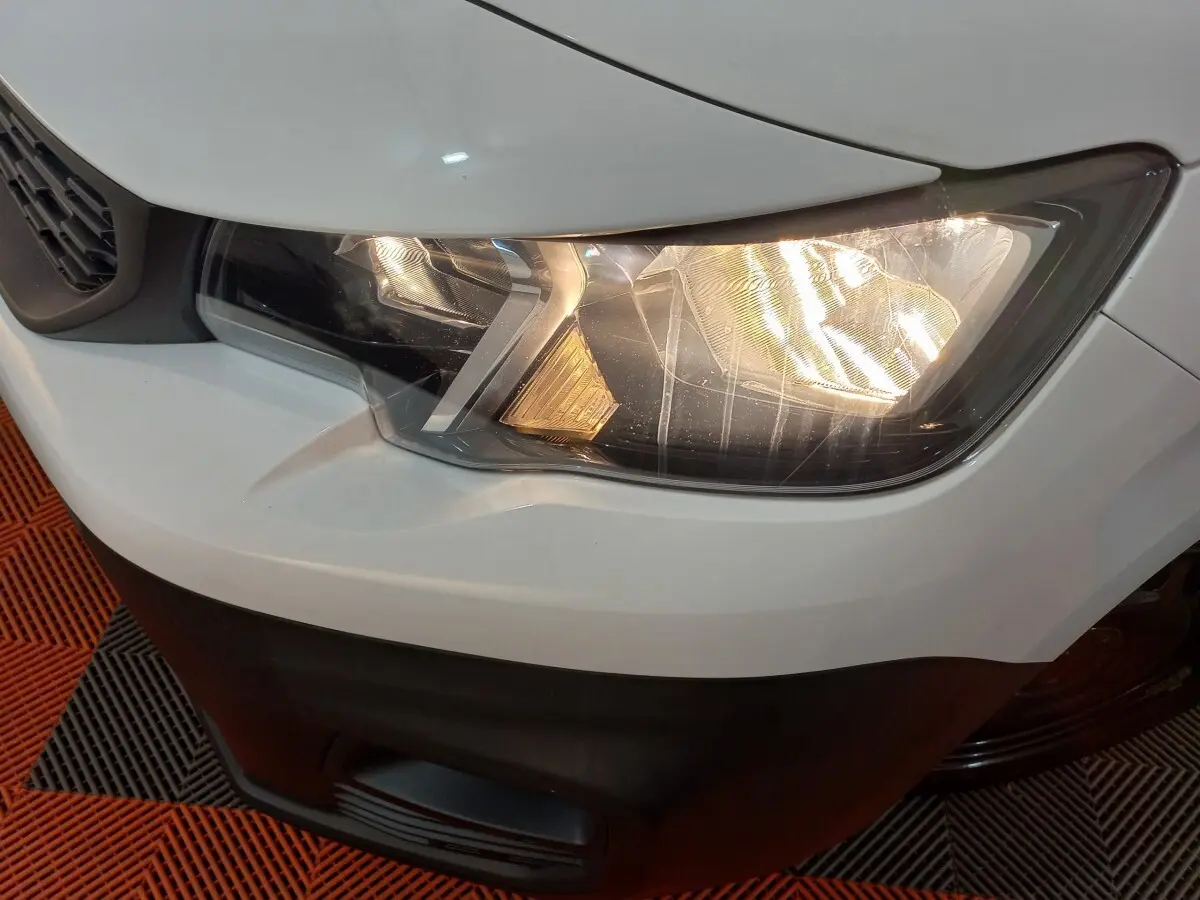 Gros plan sur le phare avant droit allumé d’un Peugeot Partner blanc, avec partie de la calandre et pare-chocs noir visible.