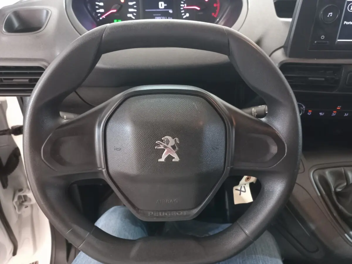 Vue rapprochée du volant noir avec logo Peugeot au centre, tableau de bord et levier de vitesses d’un Partner blanc 2021.