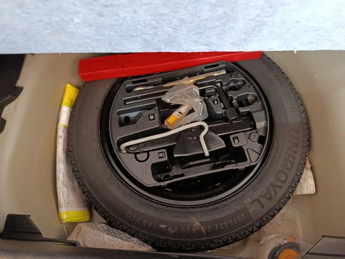 Vue plongeante sur la roue de secours noire et ses outils de bord dans le coffre d'une Peugeot 208 blanche.