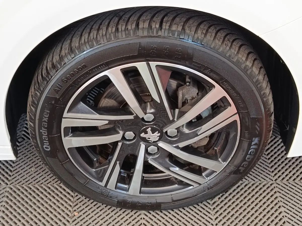 Gros plan sur la roue avant droite d'une Peugeot 208 blanche, jante alliage noire et argent avec logo Peugeot central.