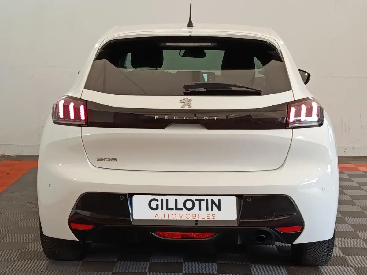 Vue arrière d'une Peugeot 208 blanche de 2021 avec feux LED allumés et logo Peugeot visible sur hayon noir.