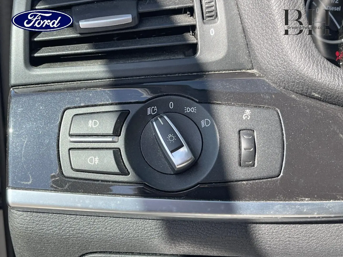 Gros plan sur la commande des phares d’un tableau de bord noir, avec logo Ford visible en haut à gauche.