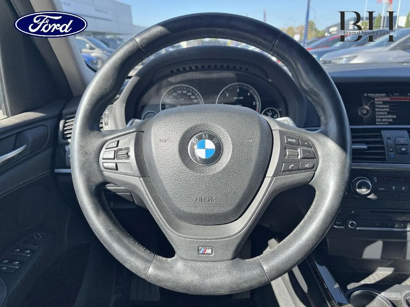 Vue rapprochée du volant cuir BMW M avec commandes intégrées, tableau de bord et écran multimédia visibles en arrière-plan.