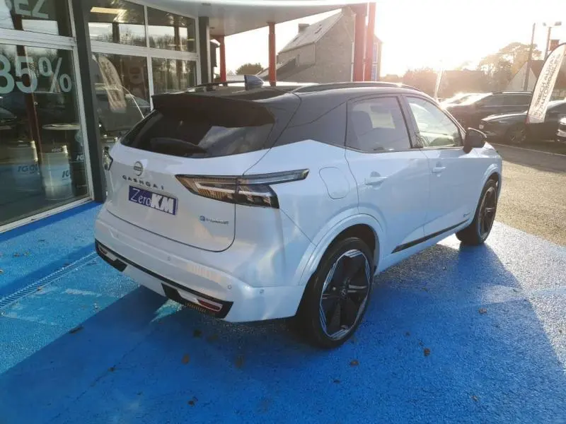 Nissan Qashqai Hybrid e-POWER blanc nacré toit noir vu en 3/4 arrière droit sur parking bleu.