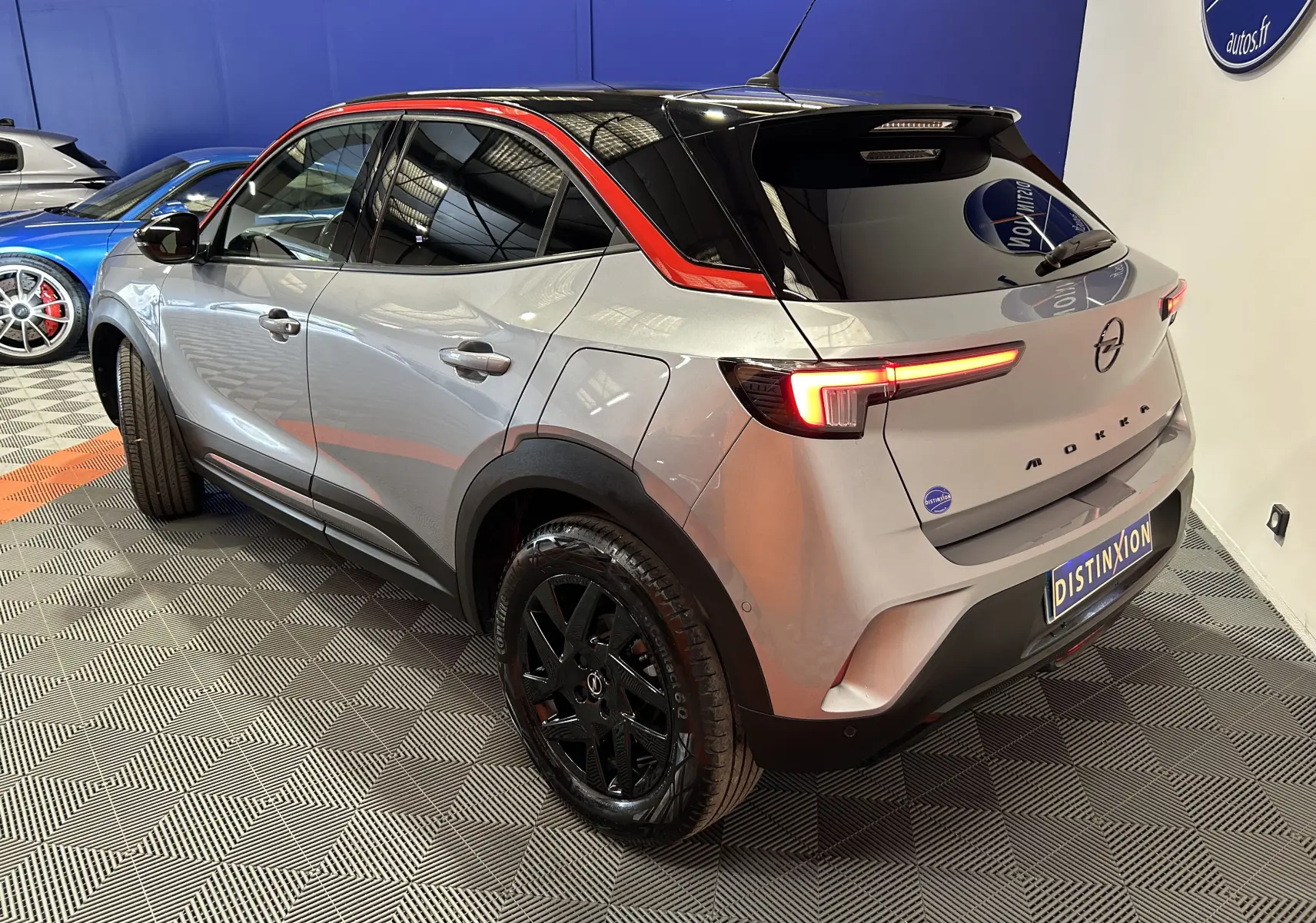 Vue 3/4 arrière droite de l’Opel Mokka gris avec toit noir et barres de toit rouges, jantes noires brillantes.