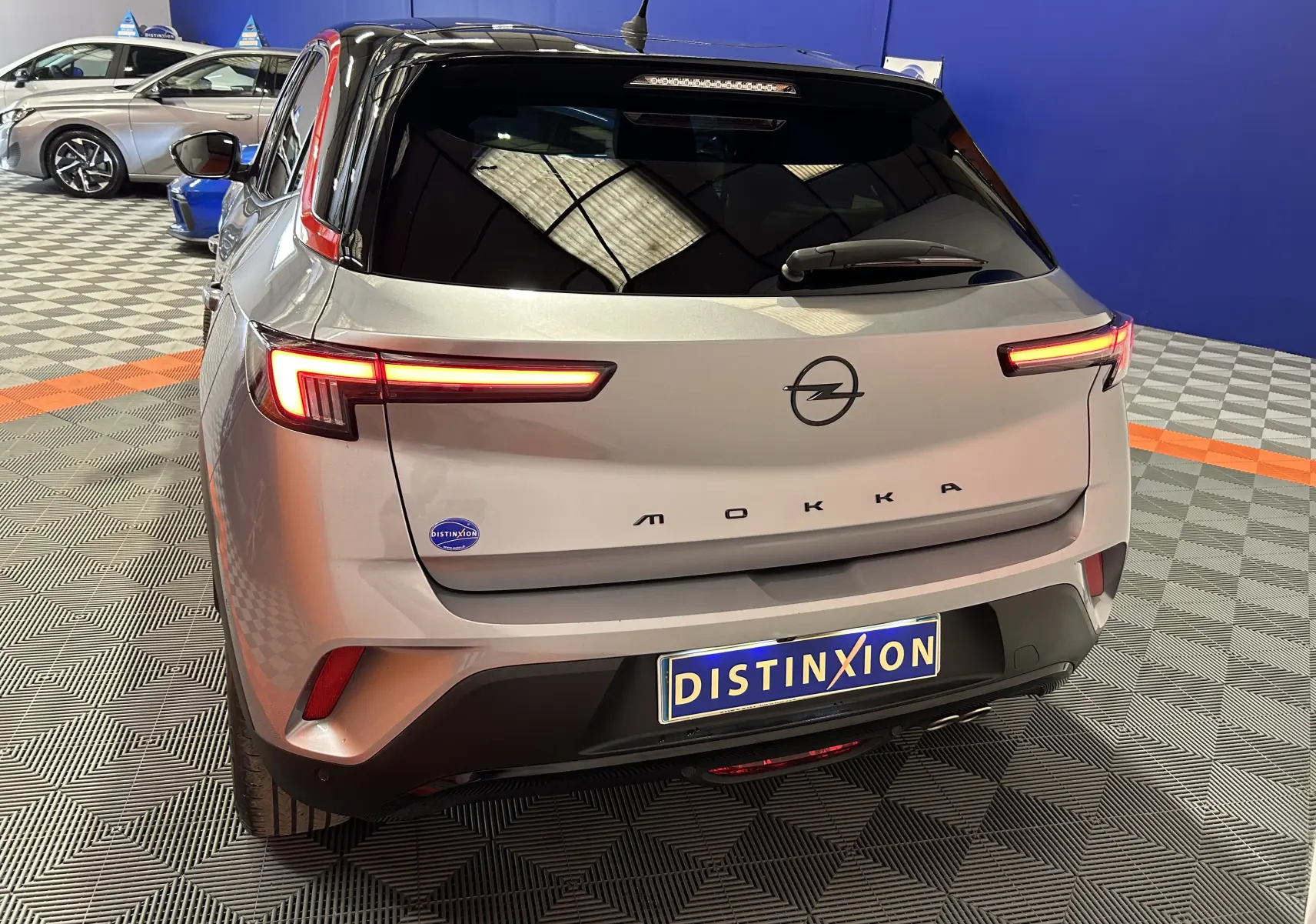 Vue 3/4 arrière d’un Opel Mokka gris avec feux arrière allumés et plaque Distinxion dans un showroom.
