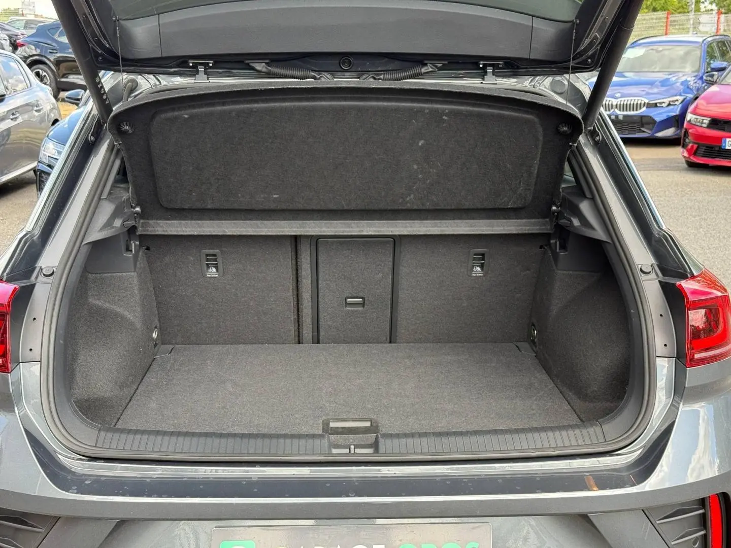 Coffre ouvert vu de l'arrière d'un Volkswagen T-Roc 2025 gris Indium métallisé avec intérieur de coffre spacieux et tapis gris.
