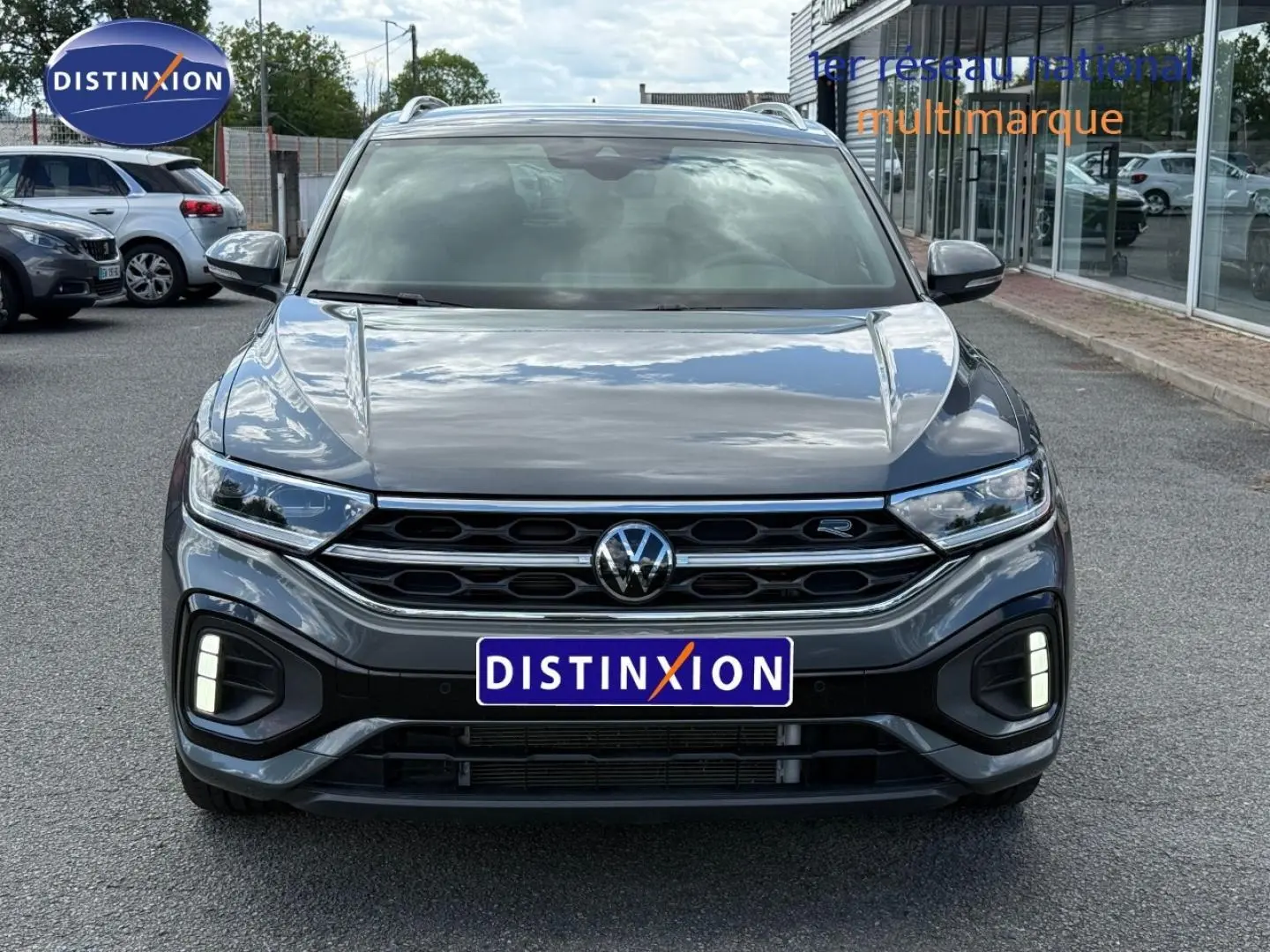 Vue avant d’un Volkswagen T-Roc gris Indium métallisé avec calandre R-Line et feux LED allumés sur un parking extérieur.