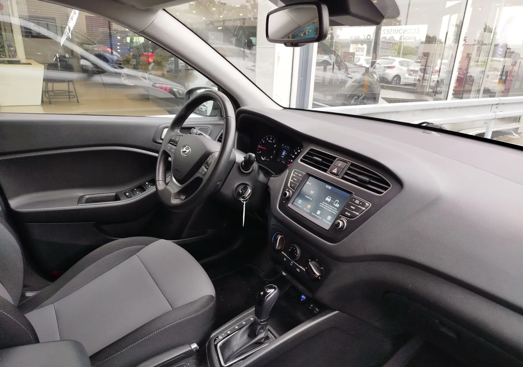 Vue intérieure côté conducteur de la Hyundai i20 2018, avec tableau de bord gris et écran tactile central.