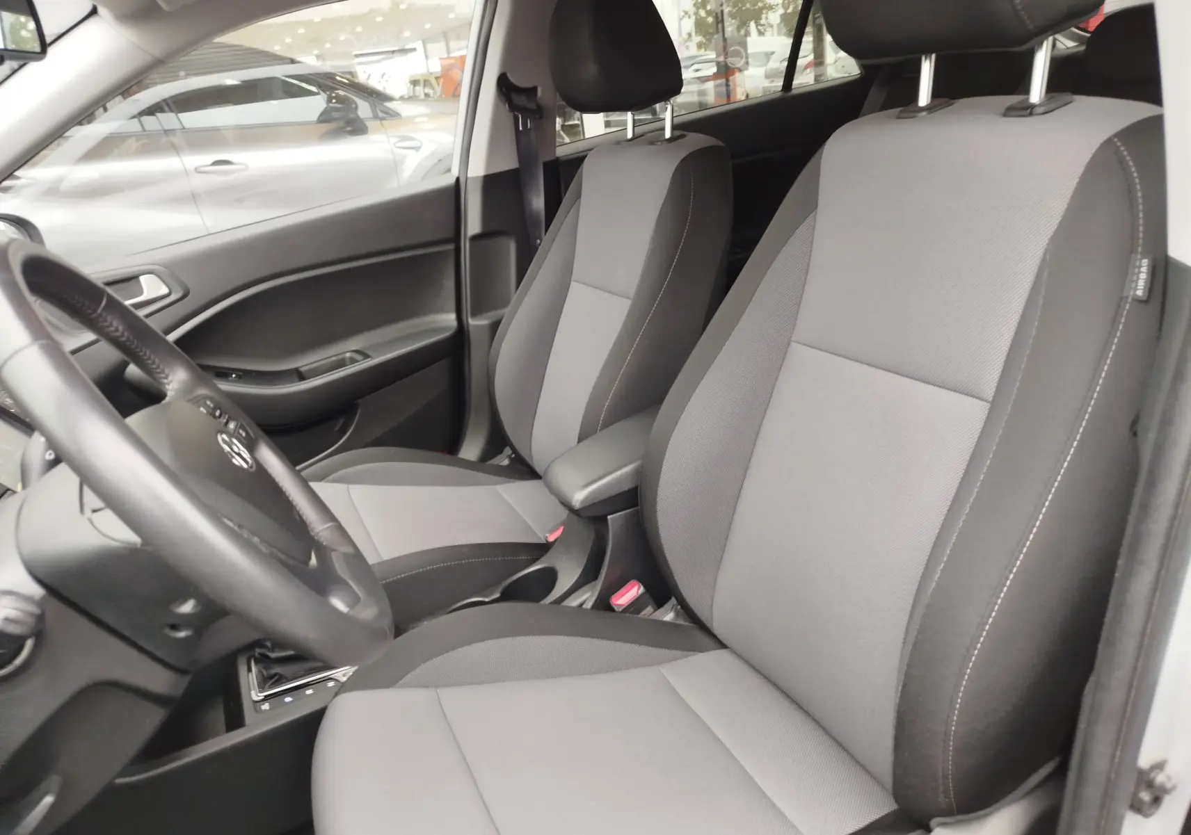 Vue intérieure côté gauche de la Hyundai i20 2018, sièges tissu gris et volant avec commandes intégrées.