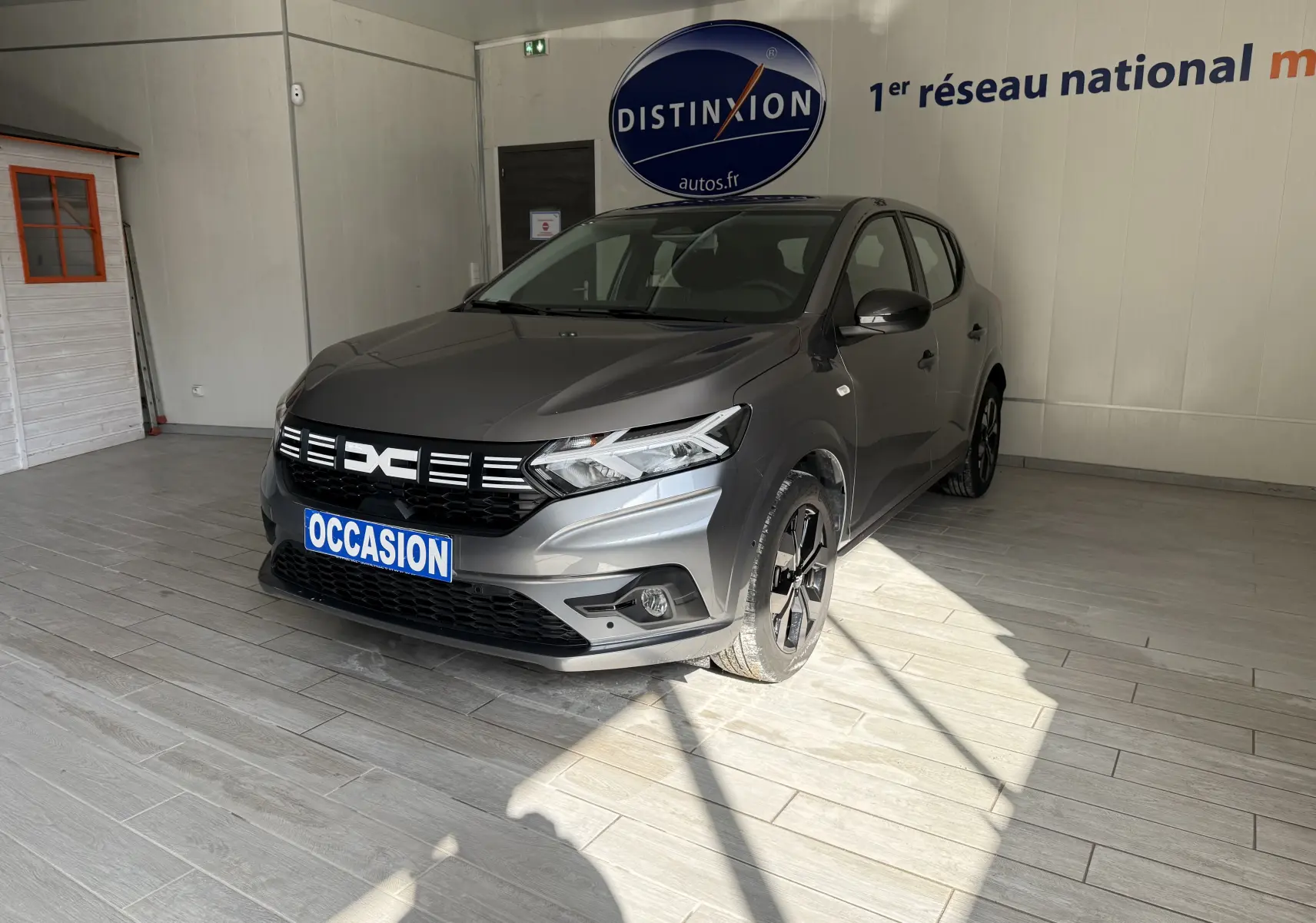 Vue 3/4 avant droite d'une Dacia Sandero gris métallisé 2025 en showroom avec plaque "OCCASION" visible.