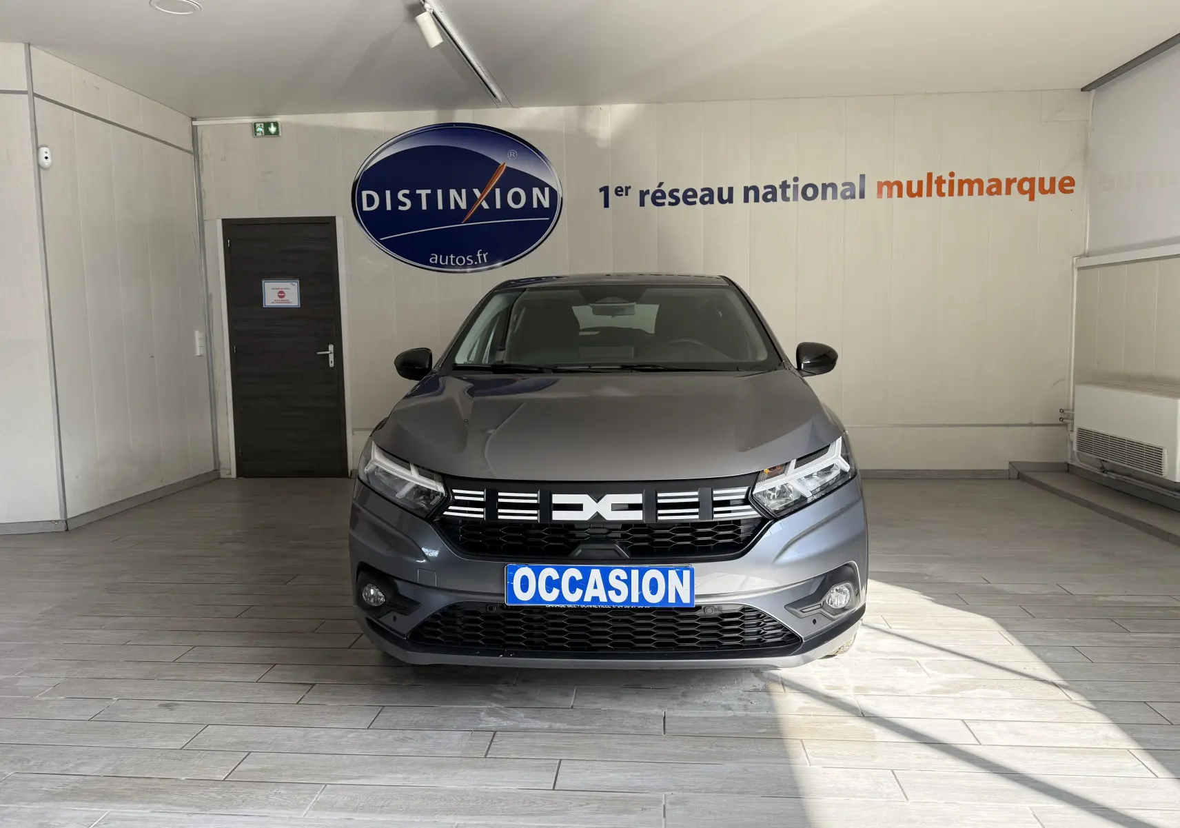 Vue frontale d'une Dacia Sandero gris métallisé 2025 en intérieur avec plaque "OCCASION" visible.