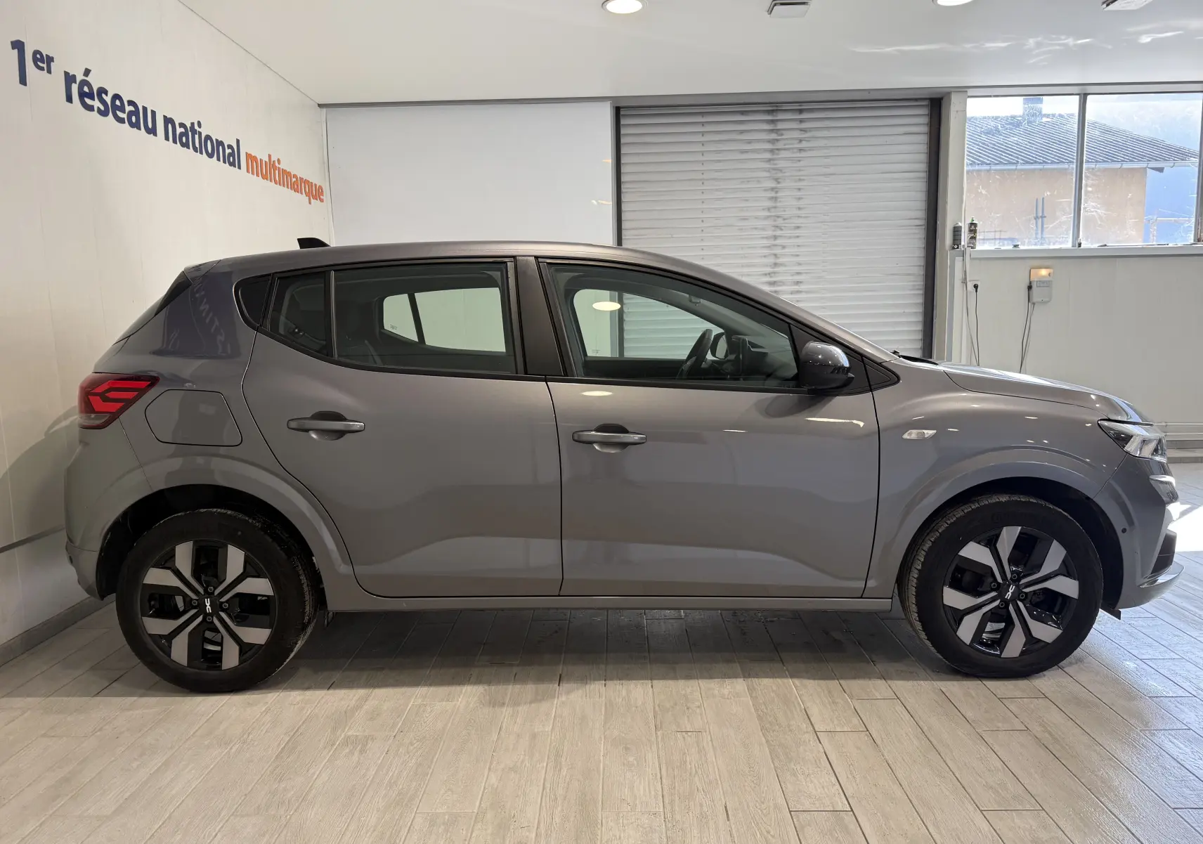 Profil droit d'une Dacia Sandero gris 2025 avec jantes alliage bicolores, garée en intérieur sur sol clair.