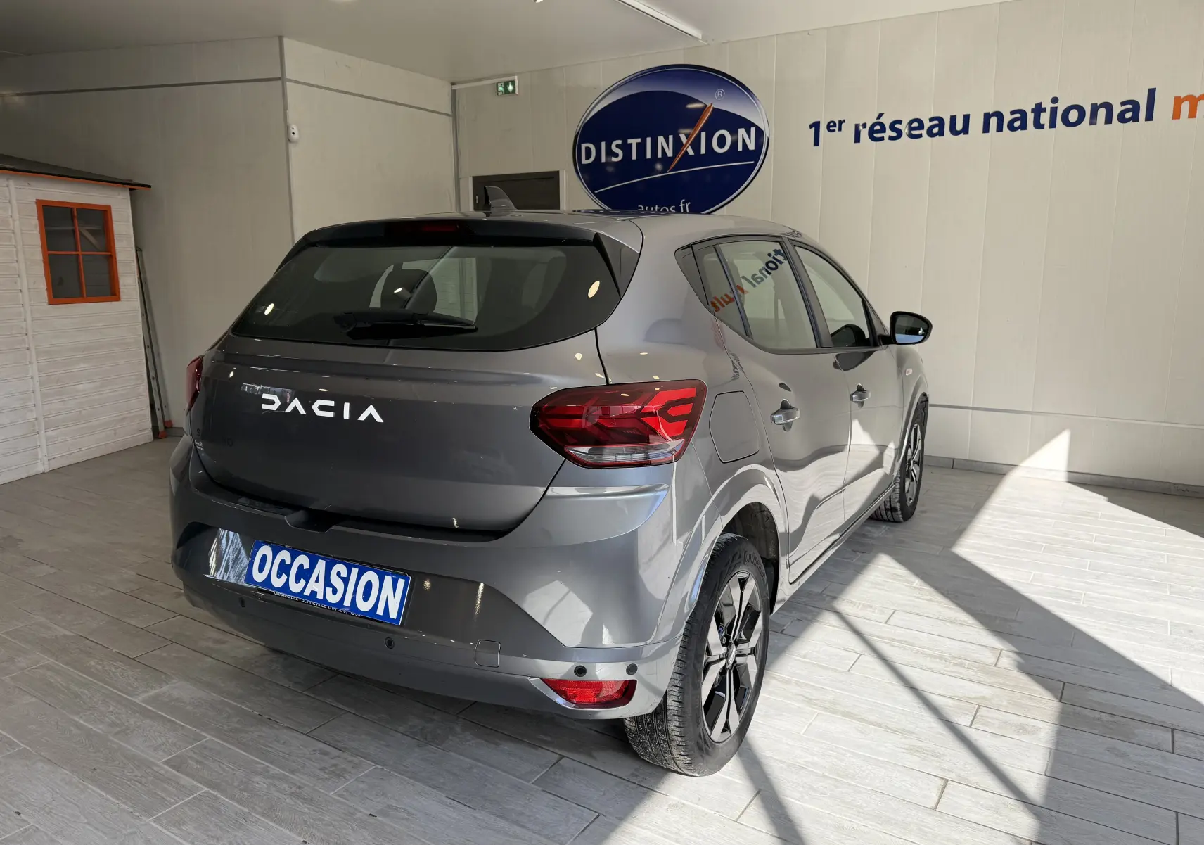Vue 3/4 arrière droite d'une Dacia Sandero gris métallisé 2025 en showroom avec plaque "OCCASION" visible.
