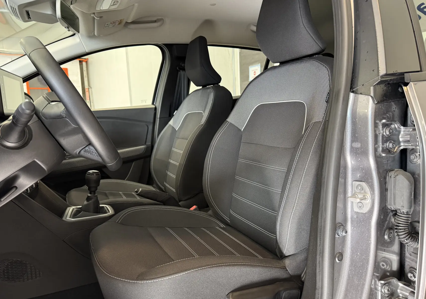 Vue intérieure côté conducteur de la Dacia Sandero gris 2025, sièges tissu noir avec surpiqûres blanches et boîte manuelle.
