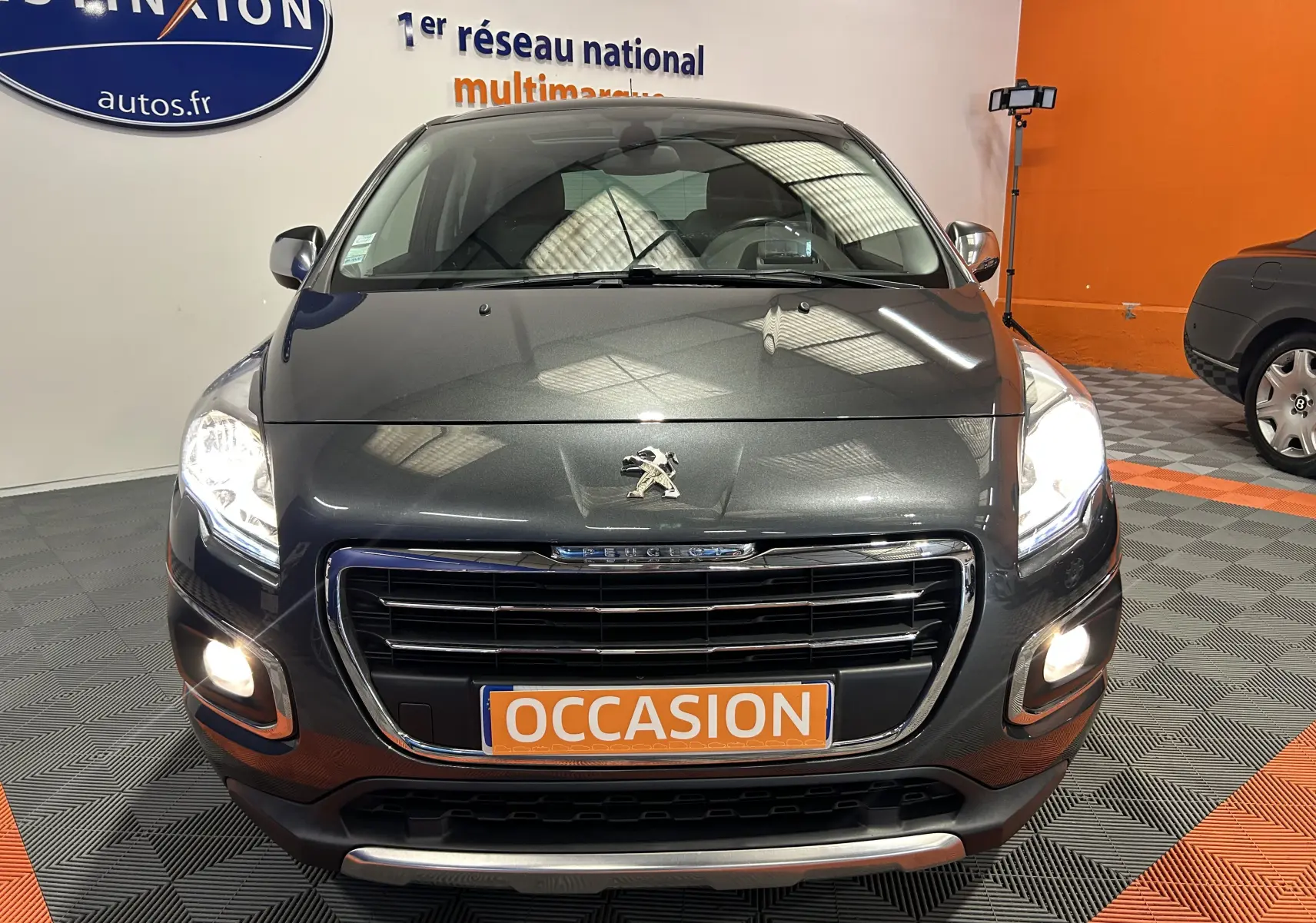 Vue avant d'un Peugeot 3008 gris avec phares allumés et plaque orange "OCCASION" en intérieur showroom.